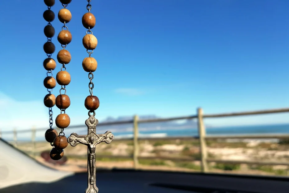 Terço de madeira com crucifixo pendurado diante de uma paisagem litorânea iluminada pelo sol, simbolizando fé e oração a Nossa Senhora Desatadora dos Nós.